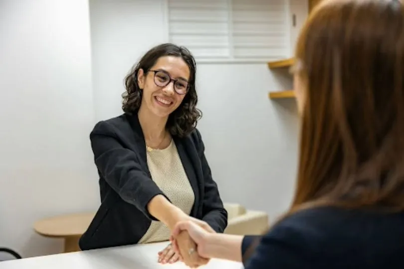 woman in glasses shakes hands across a table with another woman